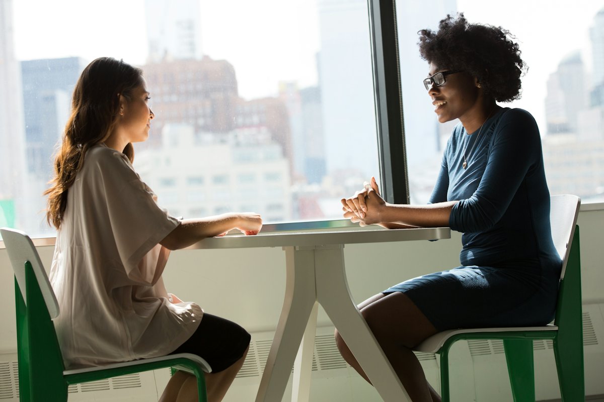 Duas mulheres conversando sentadas a uma mesa junto a uma janela ampla e iluminada